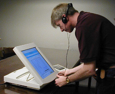Photo of man wearing headphones using a table-top machine with a large touch screen. A keypad sits nearby.