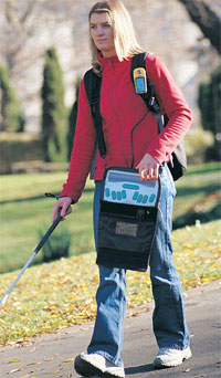 Photo of a woman walking in a park using a long cane with her right hand and reading from a BrailleNote braille display with her left hand. The GPS receiver is hooked onto the strap of her backpack.
